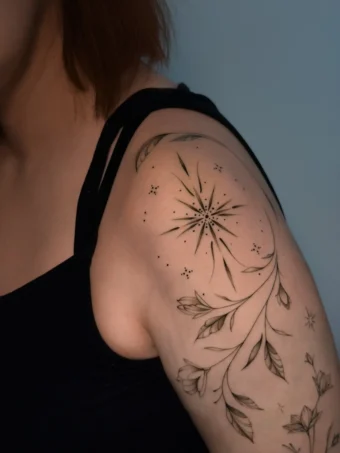 Delikatny tatuaż linework na ramieniu: gałązki z liśćmi, drobne kwiaty i gwiazda/rozbłysk z kropkami w czerni.